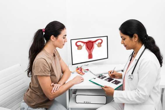 A doctor discusses ultrasound images with a patient in an office; a computer screen behind them displays a diagram of the female reproductive system.
